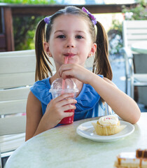 Girl sitting on cafe patio sipping bright red drink through plastic cup and nibbling cupcake