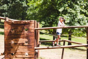 Child girl balancing on thick wooden beam at park playground using rope railings, copy space