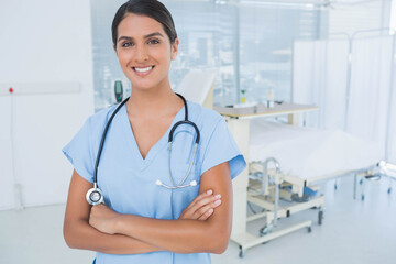 Asian woman standing with arms crossed in clinic wearing scrubs and stethoscope beside hospital bed