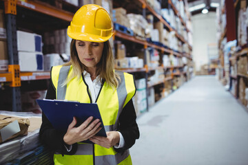 Female supervisor wearing helmet and vest standing beside pallet racks reviewing stock on clipboard