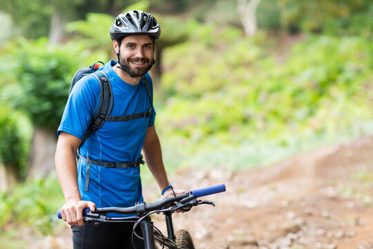 Man in 20s standing beside mountain bike on trail wearing helmet, gloves, backpack, fitness tracker - Powered by Adobe