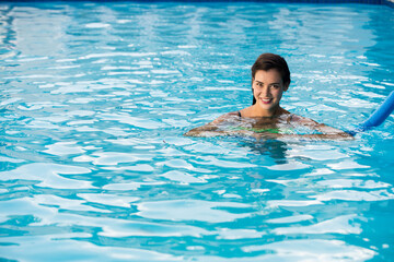 Woman in twenties floating in blue swimming pool holding blue foam pool noodle near tiled edge