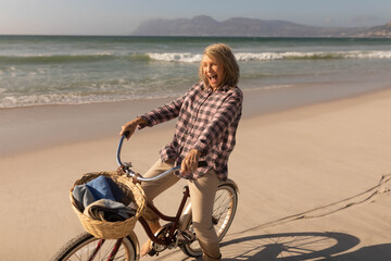 Senior woman wearing checkered shirt riding bicycle on beach with blanket in wicker basket laughing