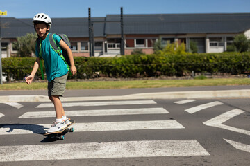 Kid is skateboarding across painted crosswalk on suburban street with bright green backpack