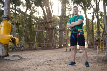 Standing man wearing harness and carabiners at forest adventure park with rope ladders, copy space