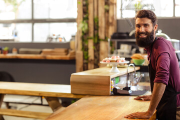 Male barista leaning on wooden counter at café, showcasing cupcakes and coffee machine, copy space