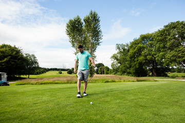 Male amateur golfer wearing glove, holding club and placing ball on tee box at parkland fairway