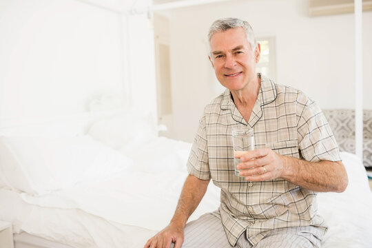 Smiling senior man sitting on bed in bright bedroom, holding glass of water, copy space