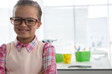 Girl standing arms crossed near window in study space, wearing glasses with plaid shirt and vest