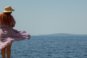Woman with summer dress and hat looking at the horizon of the Mediterranean Sea on a sunny day,...