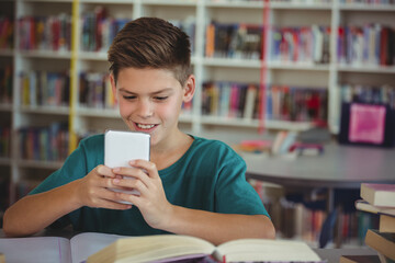 School-age boy sitting at library table holding smartphone reading open book with stacked books
