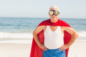 Senior white man standing on beach wearing reflective goggles and red cape, waves lapping shore