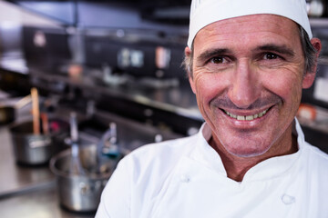 Smiling chef wearing white coat by steel countertop with pots, pans, utensils under hanging lights