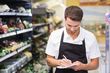 male grocery employee in apron writing notes on notepad in produce aisle, cold shelves, copy space