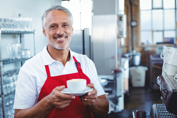 Senior male barista working behind espresso machine holding cup on saucer wearing apron in cafe