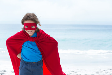 Child is standing on sandy beach wearing bright red cape and eye mask, ocean horizon visible