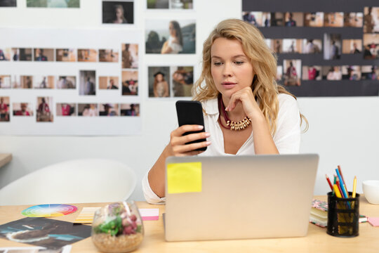 Woman sitting at wooden desk in creative studio using laptop with color wheel, sticky notes nearby - Powered by Adobe