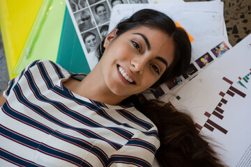 Hispanic woman lying on studio floor amid mockups, black-and-white portrait grid, charts, folders