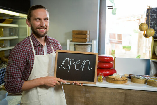 Male shopkeeper holding chalkboard sign reading open in food shop wearing white apron, copy space - Powered by Adobe