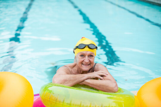 Senior woman leaning on inflatable ring in outdoor lap pool wearing yellow cap and black goggles