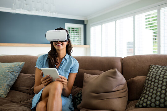 Adult Asian woman sitting on brown sofa in living room wearing white VR headset, holding tablet