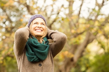 Woman standing amid autumn trees wearing cardigan sweater, wool scarf and holding knitted beanie