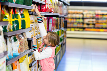 Child reaching for boxed cereal on high supermarket shelf with grains and nuts, polished tile floor