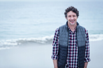 Man in his 40s wearing quilted vest standing on beach with waves rolling, copy space