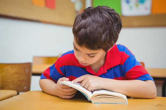 Boy sitting at wooden desk in classroom reading paperback book using finger tracking lines