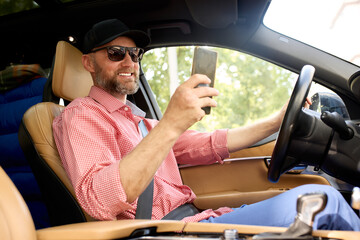 Man driving a car while holding a smartphone and smiling in sunlight