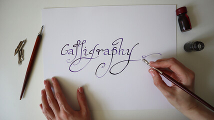 Close-up of women's hand writing word Calligraphy with ink on white paper. Brushes and an ink...