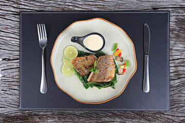 Sea bass steak served with salad greens dressed with olive oil and lemon slices on wooden tray