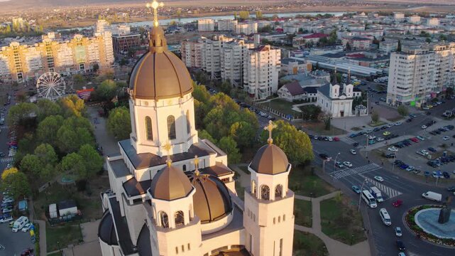 A cinematic aerial shot slowly orbits the main dome of a majestic Orthodox cathedral in Bacau, Romania, during a beautiful golden hour sunset, revealing the cityscape.