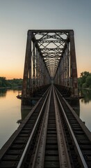 A weathered metal railway bridge stretches across a tranquil river at dawn, its intricate structure reflecting in the calm water.