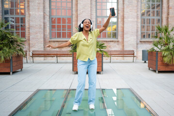 Young woman wearing headphones is dancing and singing while holding smartphone
