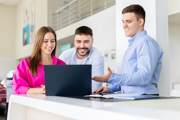 Couple with car dealer reviewing contract on laptop at dealership desk, smiling man and woman check vehicle purchase details with salesman. Lifestyle and transportation