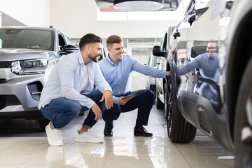 Car salesman and potential customer crouch beside sleek vehicle in modern dealership showroom. Manager holding digital tablet while explaining cars features, man attentively listening