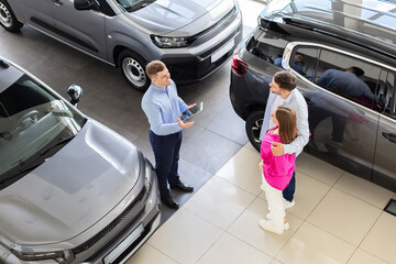 Salesman explaining vehicle features on tablet to couple at dealership, above view. Smiling man and woman standing close together listening, lifestyle and transport concept