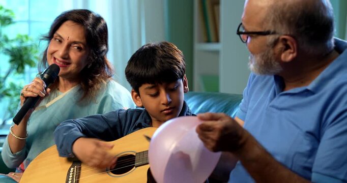 Indian grandparents with grandchild enjoying singing song together with guitar and rhythm while sitting on sofa in modern home indoors, happy family music bonding, smiling senior couple and kid