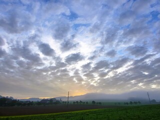 Obraz premium Scenic morning atmosphere with the sun rising over the countryside, dramatic cloud formations in the sky, lush green meadows, and gentle morning mist on the horizon
