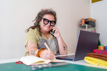 Smiling female entrepreneur managing orders taking notes on notebook and speaking on phone in her...