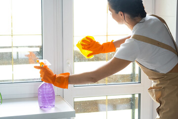 Woman in rubber gloves and apron manually washes window balcony of house with rag cleaner and mop inside interior. Restoring order and cleanliness in spring, cleaning servise