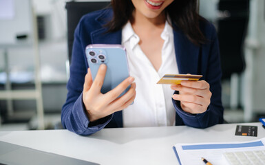 Fototapeta premium Closeup of smiling Asian businesswoman holding smartphone and credit card for online payment at office desk.