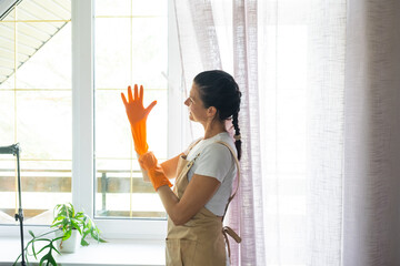 Woman in rubber gloves and apron manually washes window balcony of house with rag cleaner and mop inside interior. Restoring order and cleanliness in spring, cleaning servise