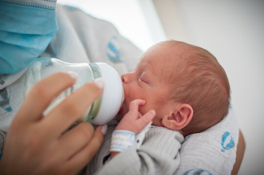 A caregiver gently feeds a newborn baby with a clear bottle in a hospital nursery. Soft lighting creates a warm atmosphere as the baby enjoys feeding time.
