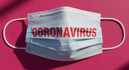 A light blue medical face mask with the word "CORONAVIRUS" in red, placed on a vibrant pink background, symbolizing the global pandemic and health crisis.