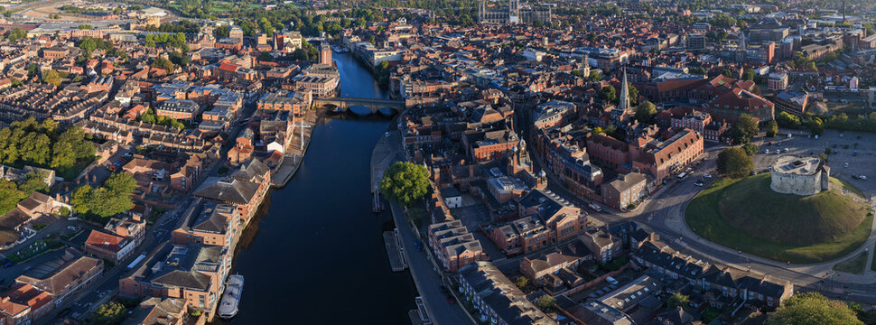 Panoramic aerial image of York showing Clifford&rsquo;s Tower, River Ouse, bridges, and dense historic rooftops at sunrise.