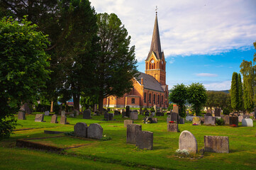 Brick church with tall steeple surrounded by a green cemetery and trees in Lillehammer, Norway. It is also known as Lillehammer kirke.