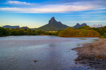 Scenic view of Rempart Mountain from Tamarin Beach, Mauritius, with calm waters in the foreground, lush greenery, and a clear sky at sunset.