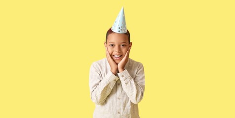 Cute African-American boy in party hat celebrating birthday on yellow background